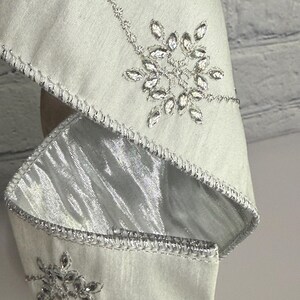 May include: Close-up of a light silver fabric with intricate silver embroidery. The design features sparkling crystal-like embellishments in a snowflake pattern. The edges are trimmed with a silver metallic border, creating a luxurious look.