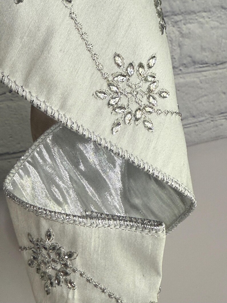May include: Close-up of a light silver fabric with intricate silver embroidery. The design features sparkling crystal-like embellishments in a snowflake pattern. The edges are trimmed with a silver metallic border, creating a luxurious look.