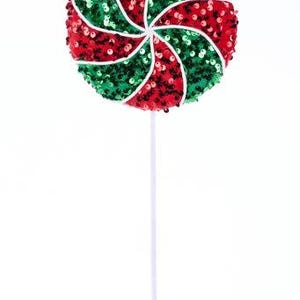 May include: A decorative lollipop on a white stick, featuring a spiral design of red and green sequins. The candy is round and has a white center, perfect for holiday decorations or festive displays.