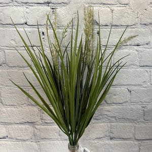 May include: A bunch of green and white artificial grass in a clear glass vase. The vase is on a white brick wall background.