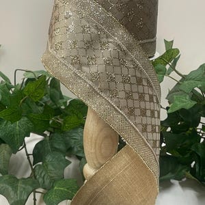 Peut inclure: Un rouleau de ruban d&eacute;coratif marron et dor&eacute;. Le ruban pr&eacute;sente un motif quadrill&eacute; avec des accents de paillettes et une bordure dor&eacute;e. Le ruban est pr&eacute;sent&eacute; sur une bobine en bois, avec un feuillage vert en arri&egrave;re-plan.