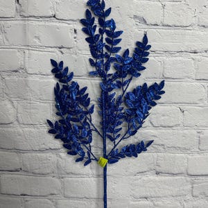 May include: A decorative artificial branch featuring multiple stems covered in vibrant blue glitter. The branch has numerous small, oval-shaped leaves extending from a central stalk, with a small yellow price tag attached near the base of the foliage.