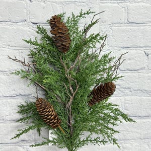 May include: A faux evergreen stem with three brown pinecones and thin, brown branches. The green foliage has a natural appearance, and the stem is set against a white brick background. This artificial plant is suitable for seasonal decor.
