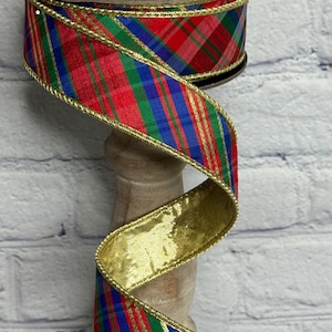 May include: A spool of wired ribbon featuring a red, green, and blue plaid pattern with metallic gold edges. The ribbon is wound around a circular cardboard core labeled d.stevens fine ribbons, displaying a shiny gold underside.