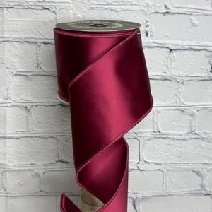 Op de afbeelding: Een spoel van diep bordeauxrood satijnen lint met een gladde, reflecterende afwerking en afgewerkte randen. Het lint is om een centrale kern gewikkeld, waarbij een enkel stuk in een zachte, vloeiende bocht naar beneden valt en de rijke kleur toont.