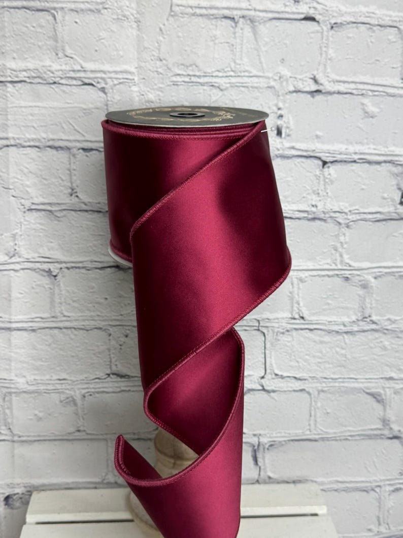 Op de afbeelding: Een spoel van diep bordeauxrood satijnen lint met een gladde, reflecterende afwerking en afgewerkte randen. Het lint is om een centrale kern gewikkeld, waarbij een enkel stuk in een zachte, vloeiende bocht naar beneden valt en de rijke kleur toont.
