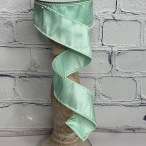 May include: A spool of mint green ribbon with a satin finish and a stitched edge. The ribbon is displayed on a decorative wooden stand against a white brick wall. The ribbon is spiraled downward.