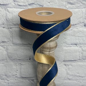 May include: A spool of double-sided ribbon featuring a deep navy blue velvet texture on one side and a smooth metallic gold finish on the other. The edges of the ribbon are lined with a thin, shimmering gold metallic trim that runs along both sides.