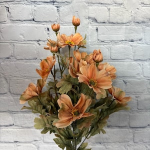 May include: A bouquet of peach-colored artificial flowers with green leaves in a clear glass vase. The flowers have a delicate, ruffled appearance and are arranged in a natural, flowing style.