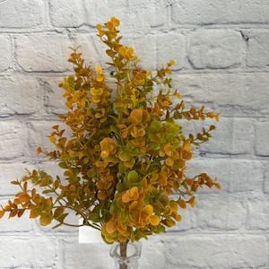 May include: A bouquet of artificial greenery with small, round, yellow-green leaves and stems. The faux plant is displayed in a clear, textured glass vase against a white brick wall background. The leaves have a gradient of colors.