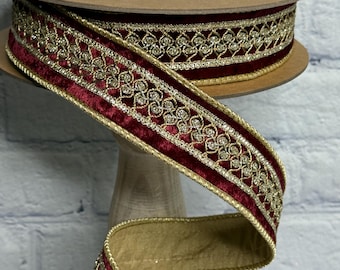 May include: A spool of burgundy velvet ribbon with gold embroidered detailing. The ribbon features a repeating pattern of gold filigree and small, sparkling accents. The edges are trimmed with gold fabric, creating a luxurious and decorative trim.