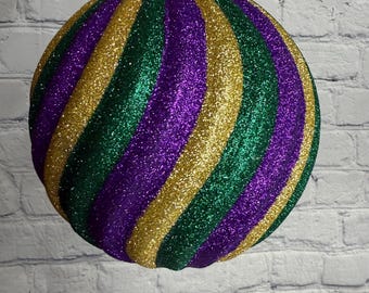 May include: A large, round Christmas ornament with a glittery finish. The ornament features alternating stripes of gold, green, and purple. It is suspended by a gold-colored cap and a thin gold string.