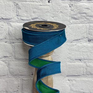 May include: A spool of double-sided ribbon featuring a deep teal blue on one side and a vibrant grass green on the other. The ribbon is wound around a black circular cardboard core with gold text that reads John Mark Enterprises.