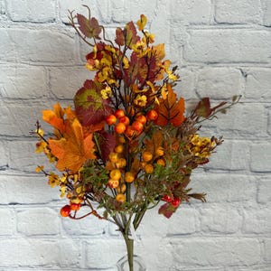 May include: A fall-themed floral arrangement featuring a variety of leaves in shades of orange, yellow, and red, along with small orange berries. The bouquet is displayed in a clear, textured glass vase, perfect for seasonal decor.
