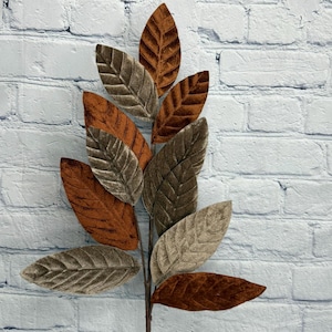 Peut inclure: Une branche artificielle décorative composée de plusieurs feuilles ovales au fini texturé semblable à du velours. Les feuilles alternent entre des tons orange brûlé profond et taupe, chacune présentant une nervure centrale marquée.