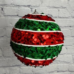 May include: A round Christmas ornament with alternating bands of red and green sequins, separated by white stripes. The ornament is suspended by a silver cap and string, against a white brick wall.