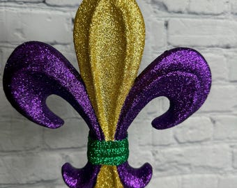 May include: A glittery fleur-de-lis ornament in gold, purple, and green. The ornament has a gold center and purple petals, with a green band at the base. It is hanging by a gold string, perfect for Mardi Gras or other festive occasions.