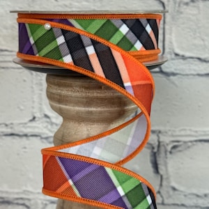 May include: A spool of decorative ribbon with a plaid pattern in orange, green, purple, black, and white. The ribbon has an orange stitched edge and is displayed on a wooden stand against a white brick background.