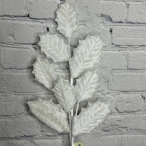 May include: A white, glitter-covered artificial holly leaf stem. The leaves have a textured appearance and are arranged along a white stem. The background is a gray brick wall. A small green tag with the word "Regency" is visible.