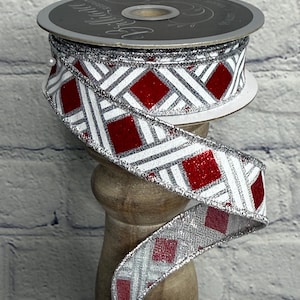 May include: A spool of decorative ribbon with a repeating pattern of red glittery squares and white crisscrossing lines, outlined in silver glitter. The ribbon is displayed on a wooden stand against a white brick wall.