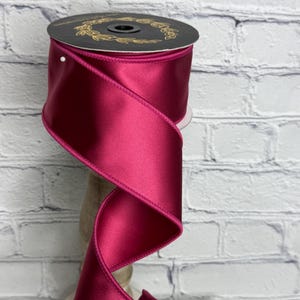 May include: A spool of deep magenta satin ribbon with a smooth, reflective finish and stitched edges. The ribbon is wound around a black circular core featuring a gold laurel wreath design and a single white pin securing the fabric in place.
