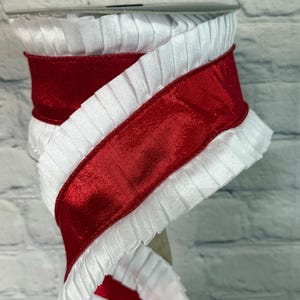 May include: A spool of white pleated ribbon with a wide, glossy red satin ribbon wrapped around it. The ribbon is likely used for crafts, gift wrapping, or decorative purposes. The white ribbon has a ruffled edge.