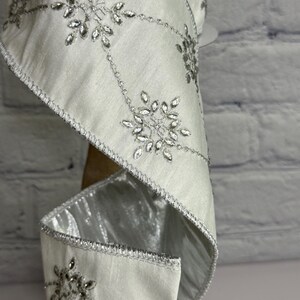 May include: White ribbon with silver snowflake embellishments. The ribbon has a satin finish and is trimmed with silver stitching. The snowflakes are made of clear, faceted crystals. This decorative ribbon is suitable for holiday and winter crafts.