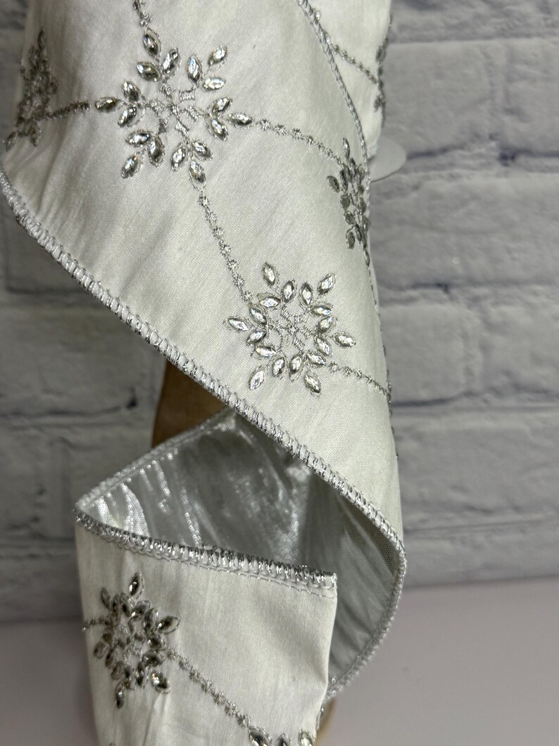 May include: White ribbon with silver snowflake embellishments. The ribbon has a satin finish and is trimmed with silver stitching. The snowflakes are made of clear, faceted crystals. This decorative ribbon is suitable for holiday and winter crafts.