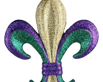 May include: Glittering fleur-de-lis ornament in gold, purple, and green. The ornament is suspended by a thin string. The design features a central gold element with purple and green accents, evoking a festive theme.