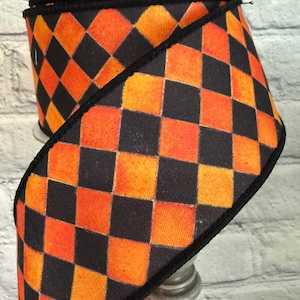 May include: A spool of decorative ribbon featuring a harlequin pattern of orange and black squares. The ribbon has a black border and is likely used for crafting or holiday decorations.