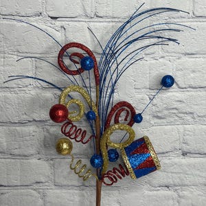 May include: A decorative floral pick featuring red, gold, and blue glittered accents. The arrangement includes spiraled wire coils, small glittered spheres, thin blue wire sprays, and a miniature drum ornament with red and blue triangular patterns.