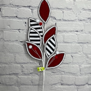 May include: A decorative floral stem featuring fabric leaves in red and black-and-white stripes. The leaves are outlined with a white stitched border and accented with small white and red pom-poms. The stem is white.