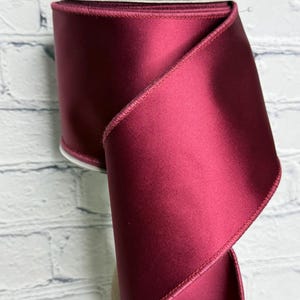 Op de afbeelding: Een brede rol dieprood satijnen lint met een gladde, reflecterende afwerking en gestikte randen. Het lint is om een centrale kern gewikkeld, waarbij een deel van de stof in een zachte, elegante bocht naar beneden valt en de rijke kleur toont.