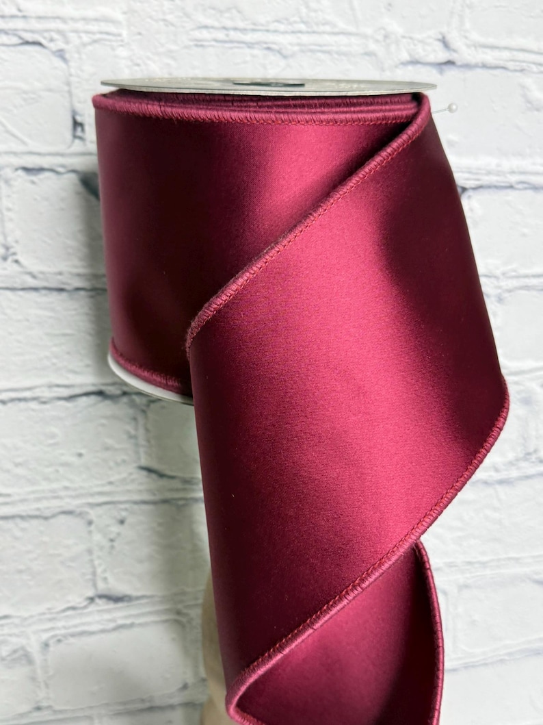 Op de afbeelding: Een brede rol dieprood satijnen lint met een gladde, reflecterende afwerking en gestikte randen. Het lint is om een centrale kern gewikkeld, waarbij een deel van de stof in een zachte, elegante bocht naar beneden valt en de rijke kleur toont.