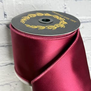 Op de afbeelding: Een brede rol dieprood satijnen lint met een gladde, glanzende afwerking en gestikte randen. De bovenkant van de rol is voorzien van een zwart rond label met een gouden lauwerkransontwerp en de tekst John Mark Enterprises erop gedrukt.