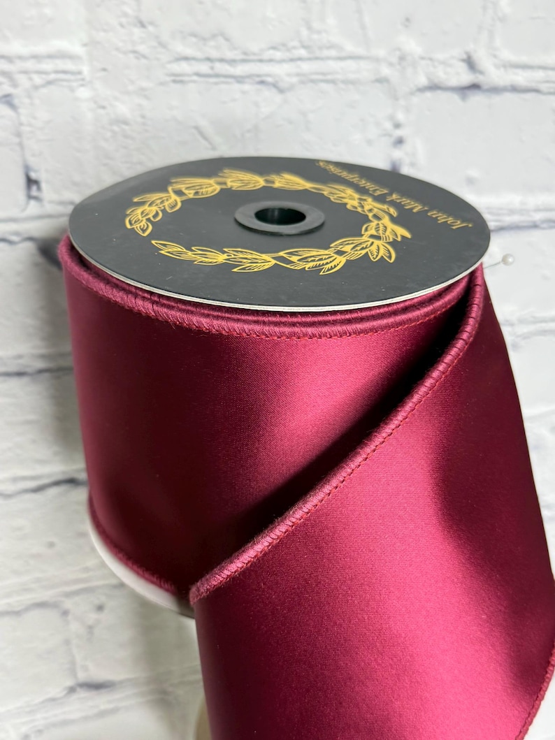 Op de afbeelding: Een brede rol dieprood satijnen lint met een gladde, glanzende afwerking en gestikte randen. De bovenkant van de rol is voorzien van een zwart rond label met een gouden lauwerkransontwerp en de tekst John Mark Enterprises erop gedrukt.