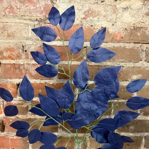 May include: A bouquet of artificial leaves in a clear glass vase. The leaves are a deep navy blue, with a slightly textured appearance. The stems are a light green color. The background is a brick wall.