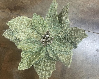 Op de afbeelding: Een grote, getextureerde, groene poinsettia bloem met een glinsterend centrum. De bloem heeft meerdere lagen bloemblaadjes en is waarschijnlijk een decoratief item voor de feestdagen. De bloemblaadjes hebben een geweven uiterlijk.