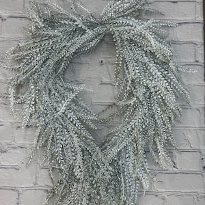 May include: A teardrop-shaped wreath constructed from artificial fern-like foliage. The leaves are a muted sage green color with a heavy dusting of white, frosted-looking powder, creating a textured, layered appearance throughout the entire piece.