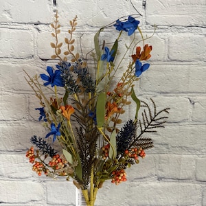May include: A bouquet of artificial flowers with blue and orange flowers, green leaves, and brown stems. The bouquet is against a white brick wall.