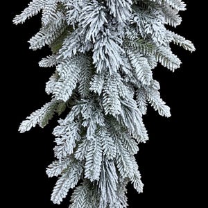 May include: A teardrop-shaped artificial evergreen swag featuring dense, needle-like foliage heavily coated in a thick layer of white, snow-like flocking. The branches are arranged in a downward-tapering cluster with visible green needles.