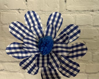 May include: A blue and white gingham paper flower with a blue pom-pom center. The flower is on a white brick background.
