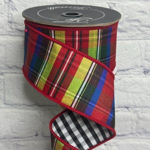 May include: A spool of wired ribbon featuring a vibrant multi-colored plaid pattern with red, yellow, green, and blue stripes on one side and a black and white gingham check pattern on the reverse, finished with a solid red stitched edge.