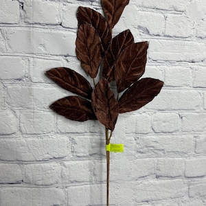 May include: A decorative artificial plant stem featuring multiple dark brown velvet leaves with visible veining. The stem is thin and brown, and a small, bright yellow rectangular tag is attached to the lower portion of the main stem.
