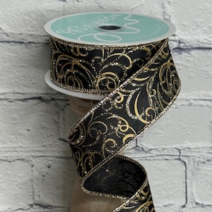 May include: A spool of black ribbon featuring a repeating gold glitter swirl pattern. The ribbon has metallic gold finished edges and is wrapped around a circular spool with a light blue label that reads RG RIBBON in white text.