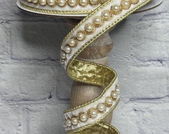 May include: A spool of white ribbon featuring a central row of cream-colored faux pearls bordered by metallic gold trim. The ribbon has a textured white fabric base and a shimmering gold underside, wound around a dark circular spool top.