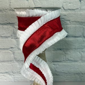 May include: A spool of decorative ribbon featuring a vibrant red velvet center bordered by ruffled white fabric. The ribbon is displayed on a neutral-toned spool, against a white brick background. This ribbon is suitable for crafts and gift wrapping.