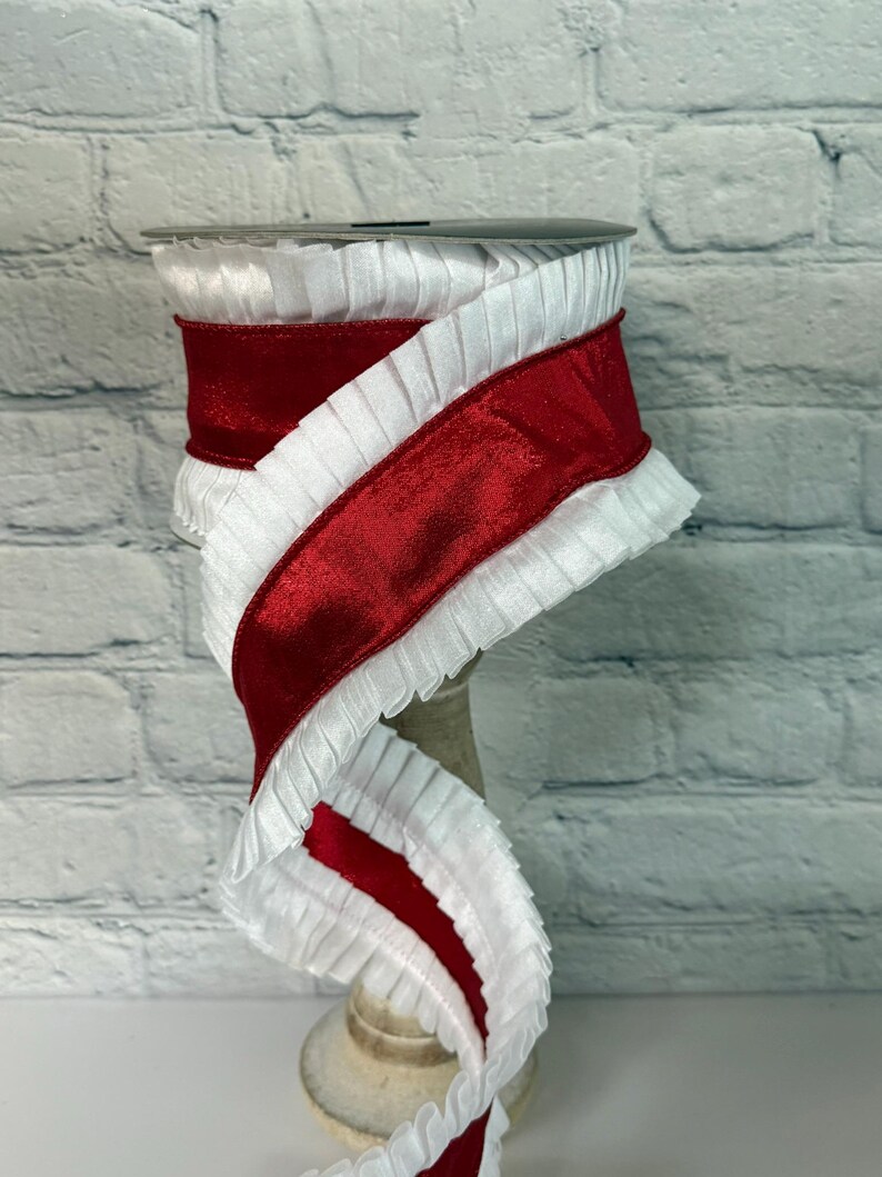May include: A spool of decorative ribbon featuring a vibrant red velvet center bordered by ruffled white fabric. The ribbon is displayed on a neutral-toned spool, against a white brick background. This ribbon is suitable for crafts and gift wrapping.