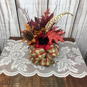 May include: A fall-themed centerpiece with red, brown, and gold leaves, a pine cone, and a red and green plaid bow. The centerpiece is on a white lace table runner.