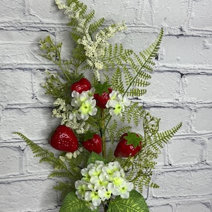 May include: A faux floral stem featuring bright red strawberries, white flowers, and green fern-like foliage. The artificial arrangement is set against a white brick background, creating a fresh and vibrant aesthetic. Perfect for home decor or crafting.